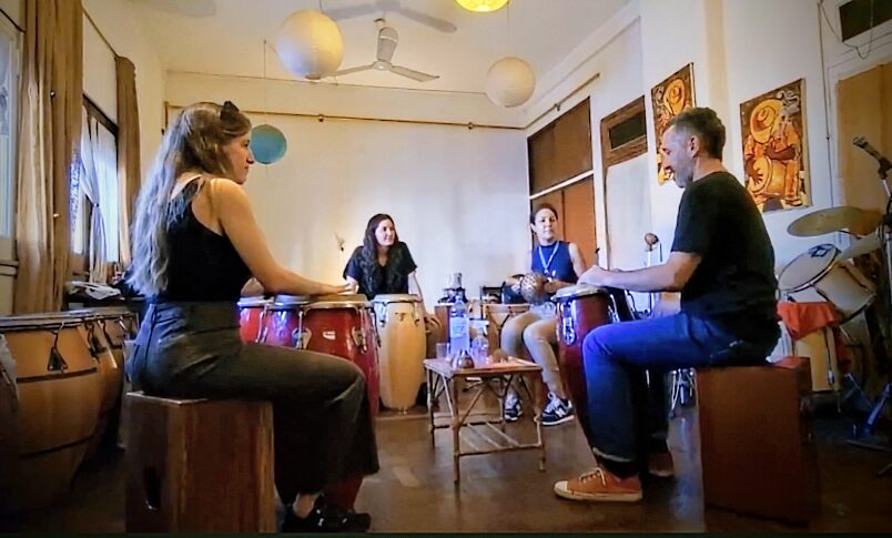 Group of people sitting in a circle on box drums with latin drums between their legs in a room full of drums